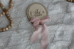 Wooden She’s Here baby announcement sign with pink chiffon bow on a cozy knit blanket, styled with wooden bead decor.