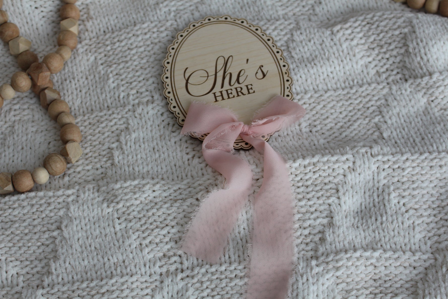 Wooden She’s Here baby announcement sign with pink chiffon bow on a cozy knit blanket, styled with wooden bead decor.