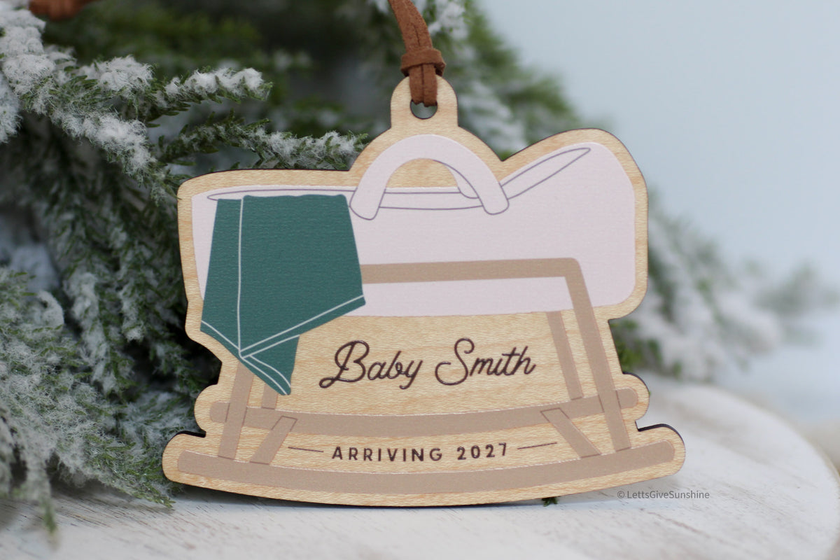 Personalized pregnancy announcement Christmas ornament featuring a wooden rocking baby cradle with a green blanket, custom Baby Smith name, and Arriving 2027, perfect for a holiday pregnancy reveal.
