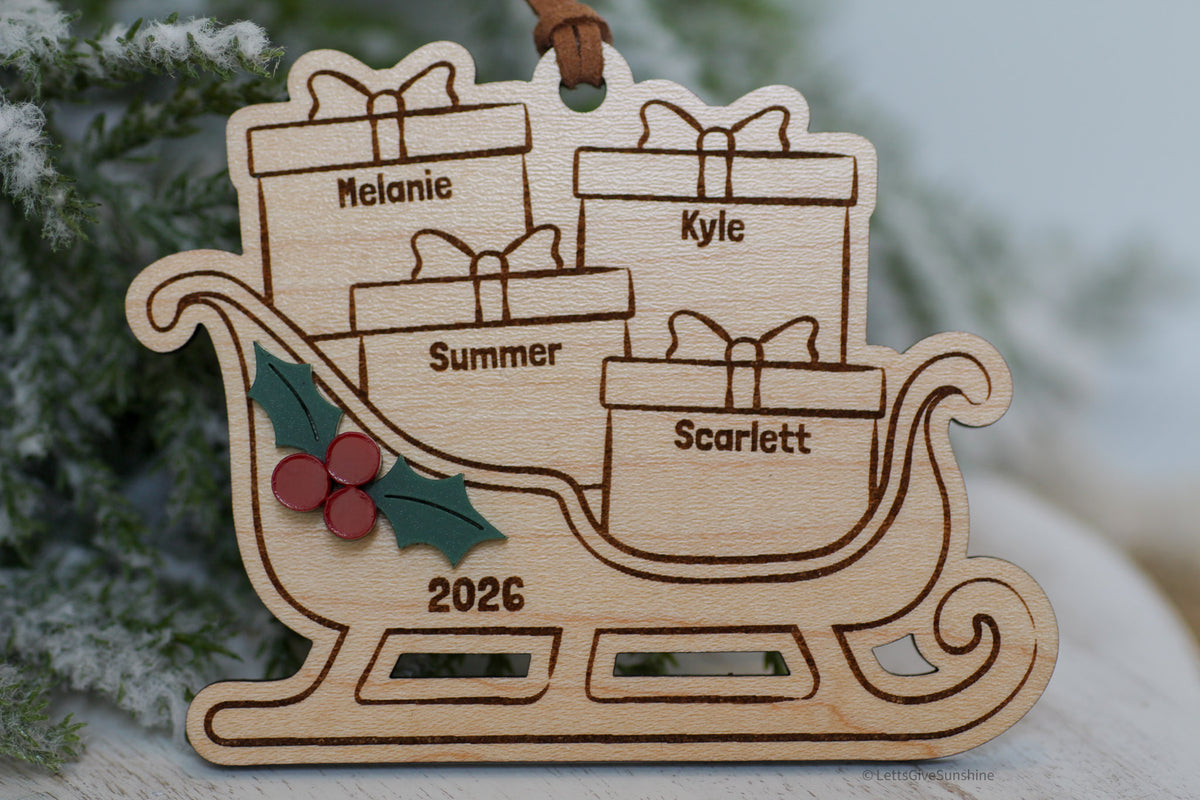 A personalized wooden Christmas ornament shaped like a sleigh carrying four gift boxes, with engraved names Melanie, Kyle, Summer, and Scarlett, the year 2026, and small holly leaves and berries. It hangs against a softly blurred festive background.