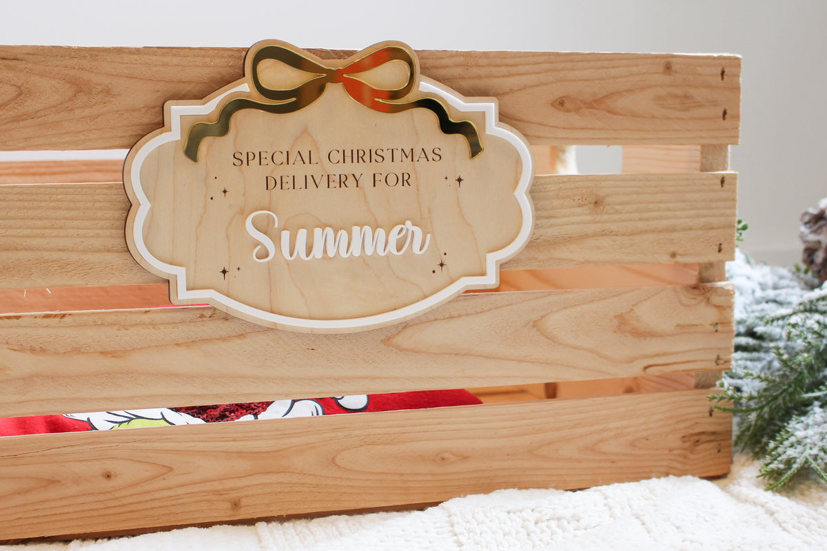 Personalized wooden Christmas Eve box plaque attached to a wooden crate, featuring the name Summer in white acrylic lettering, a gold bow and engraved details.