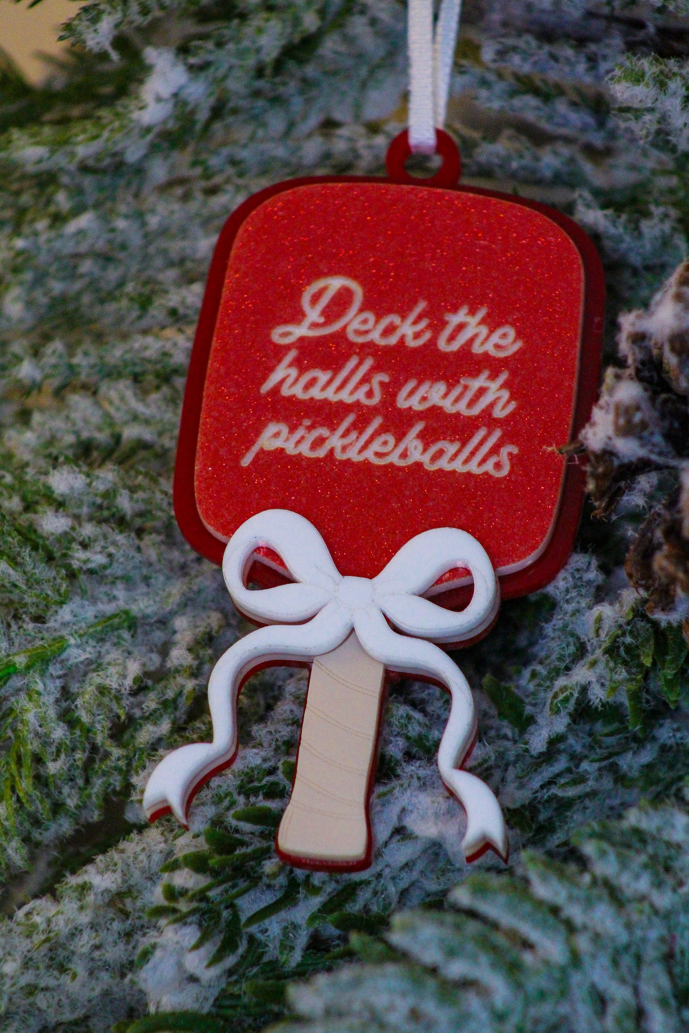Red glitter pickleball paddle Christmas ornament with a layered white bow and the words Deck the halls with pickleballs.