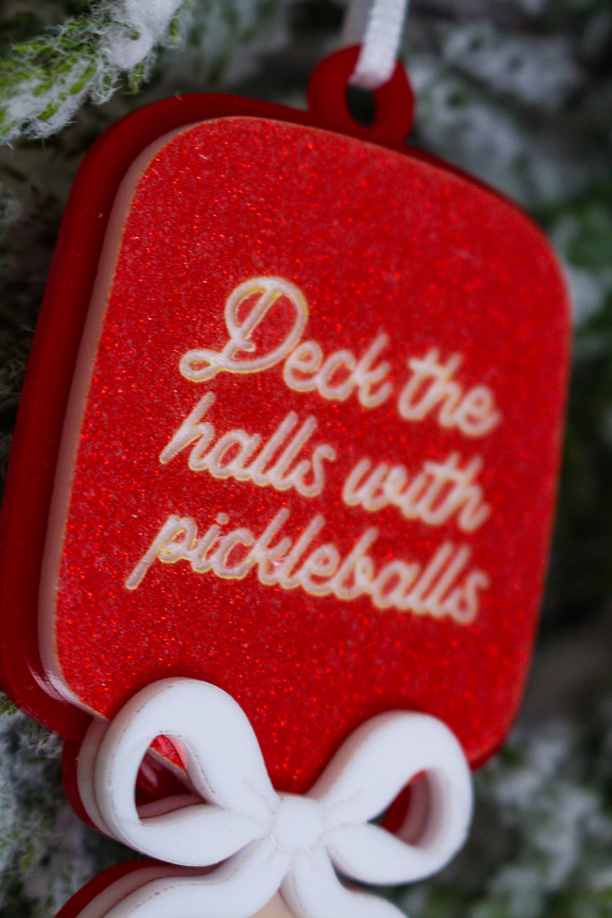 Red glitter pickleball paddle Christmas ornament with a layered white bow and the words Deck the halls with pickleballs.