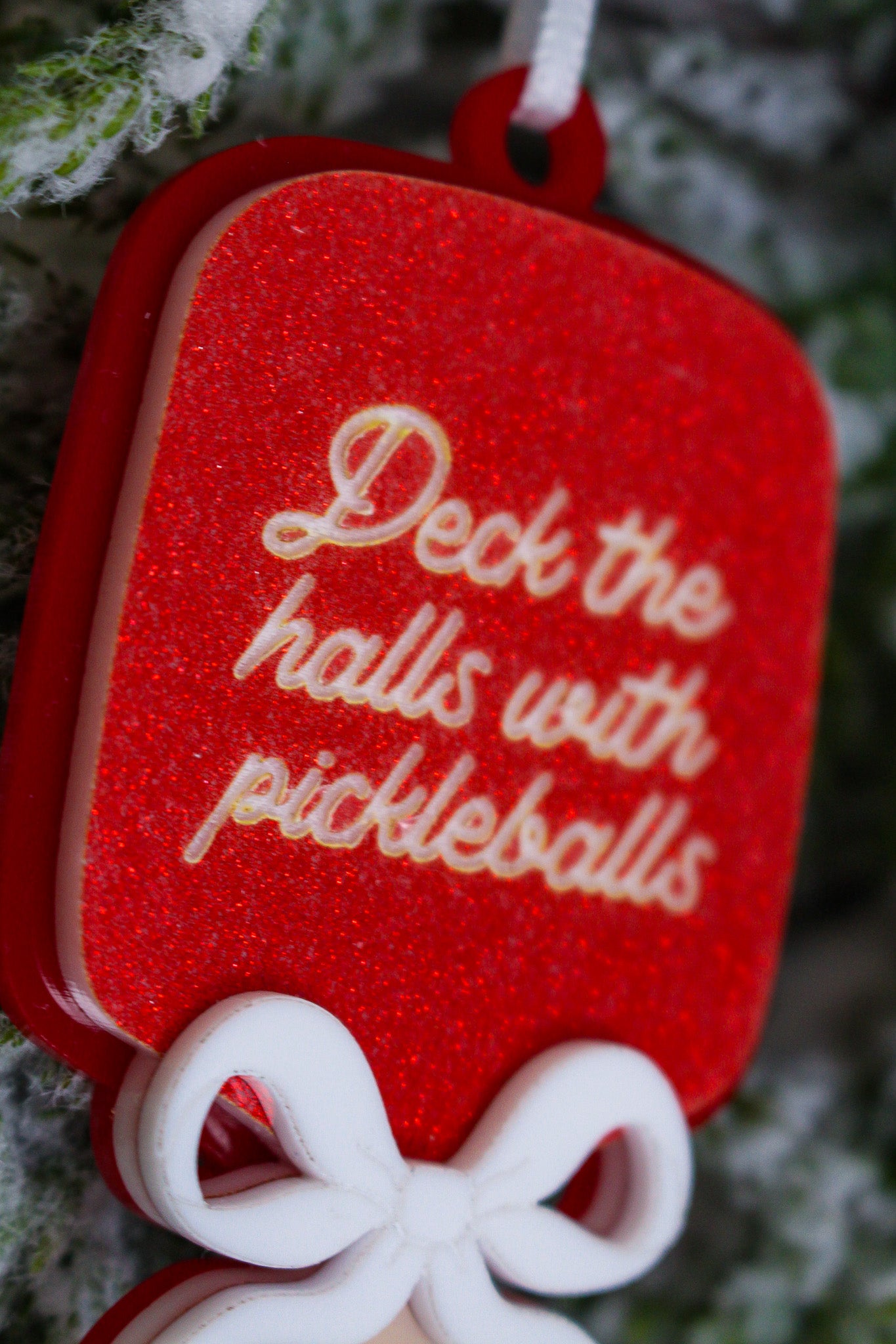 Red glitter pickleball paddle Christmas ornament with a layered white bow and the words Deck the halls with pickleballs.