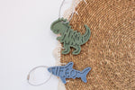 Personalized green dinosaur and blue shark bag tags with the names Tyson and Theo, attached to a woven backpack with silver cable loops.