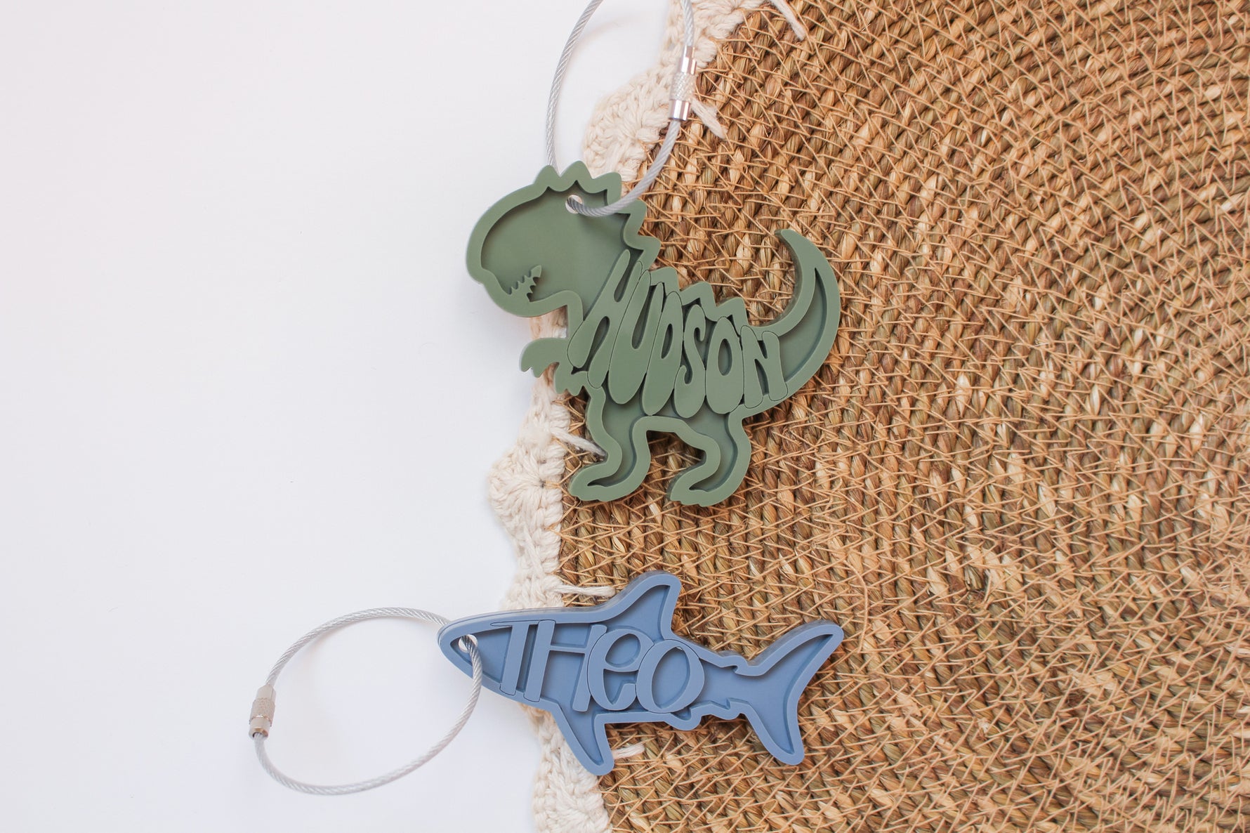 Personalized green dinosaur and blue shark bag tags with the names Tyson and Theo, attached to a woven backpack with silver cable loops.