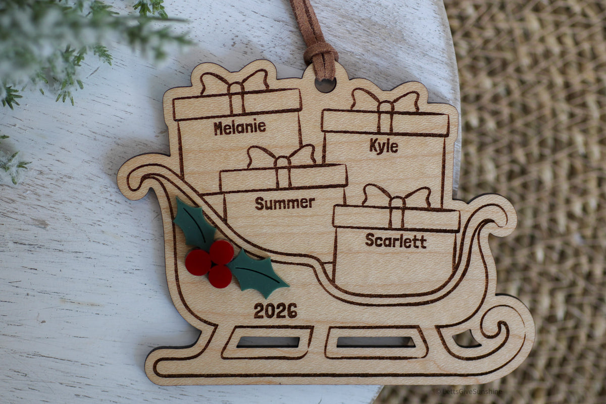 Custom family Christmas ornament 2026 featuring a wooden Santa sleigh filled with engraved name presents, layered holly berries, and a brown hanging ribbon.