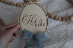 Wooden He’s Here baby announcement sign with blue fabric bow, held over a soft knit blanket with wooden bead decor.