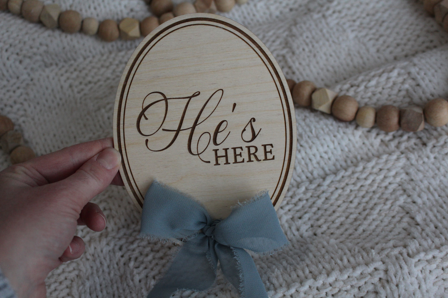 Wooden He’s Here baby announcement sign with blue fabric bow, held over a soft knit blanket with wooden bead decor.