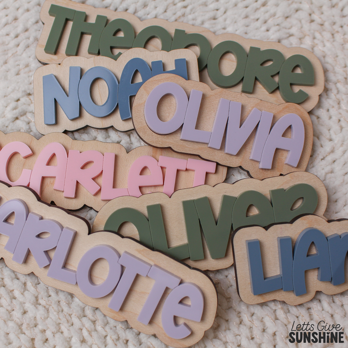 Close-up of interchangeable personalized wooden name pieces with raised acrylic lettering in pink, blue, purple and green, designed for a reusable first day of daycare dry erase milestone board.