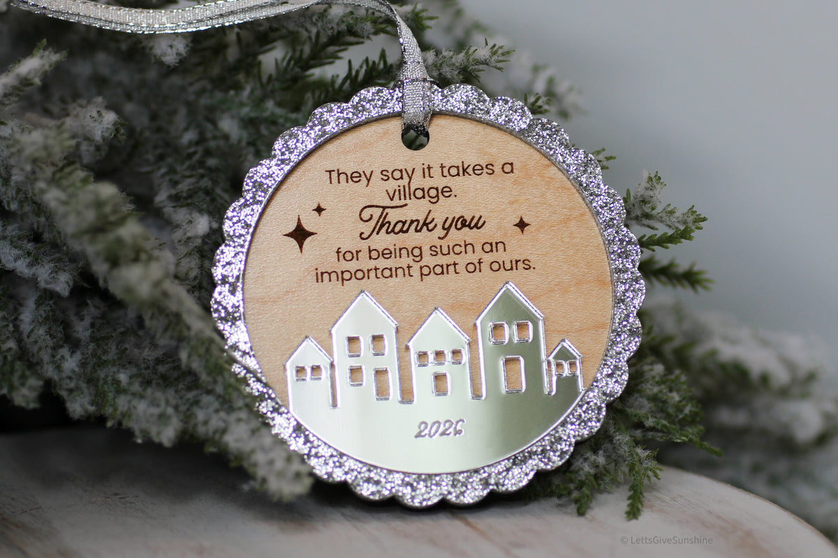 It Takes a Village 2026 Christmas ornament with engraved thank you message, silver glitter border and mirrored acrylic houses hanging on a snowy Christmas tree.