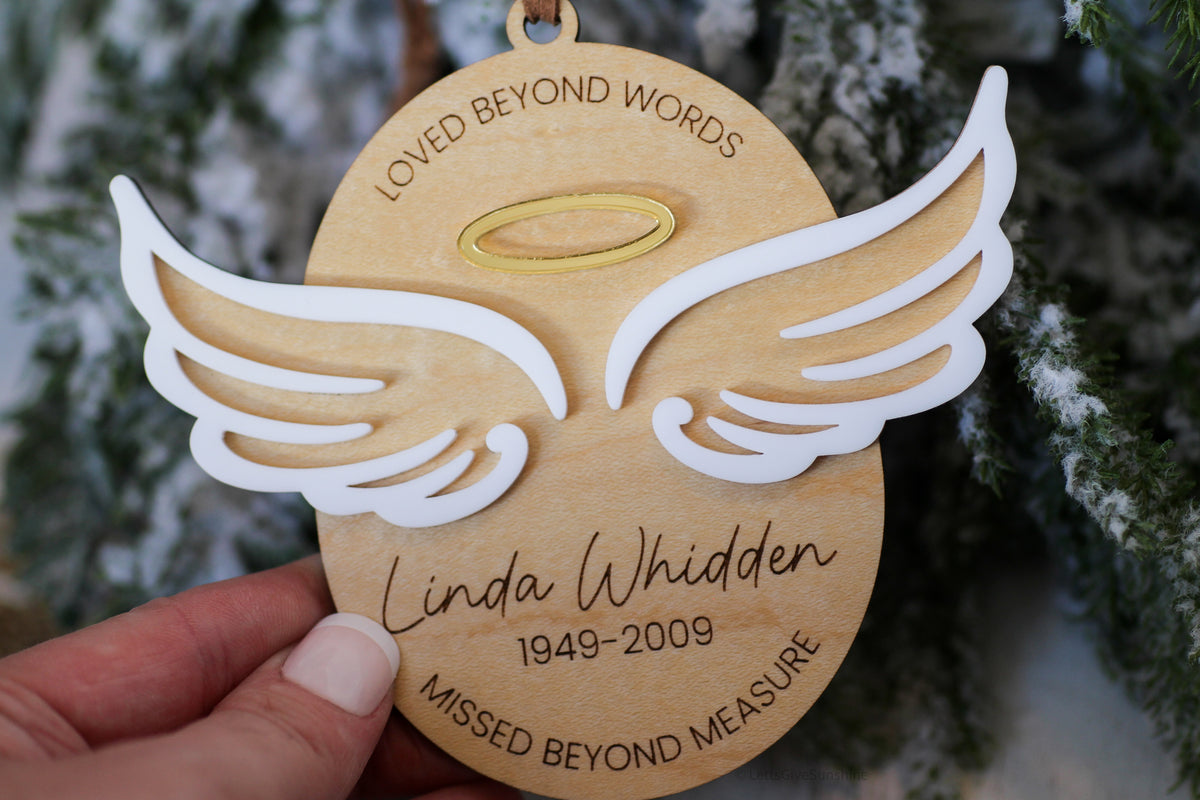 Personalized bereavement ornament with angel wings and halo, featuring the name Linda Whidden, memorial years, and a decorative wood oval shape