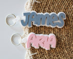 Two personalized layered acrylic name bag tags featuring James in blue and Aria in pink, each with a white backing and silver keyring.