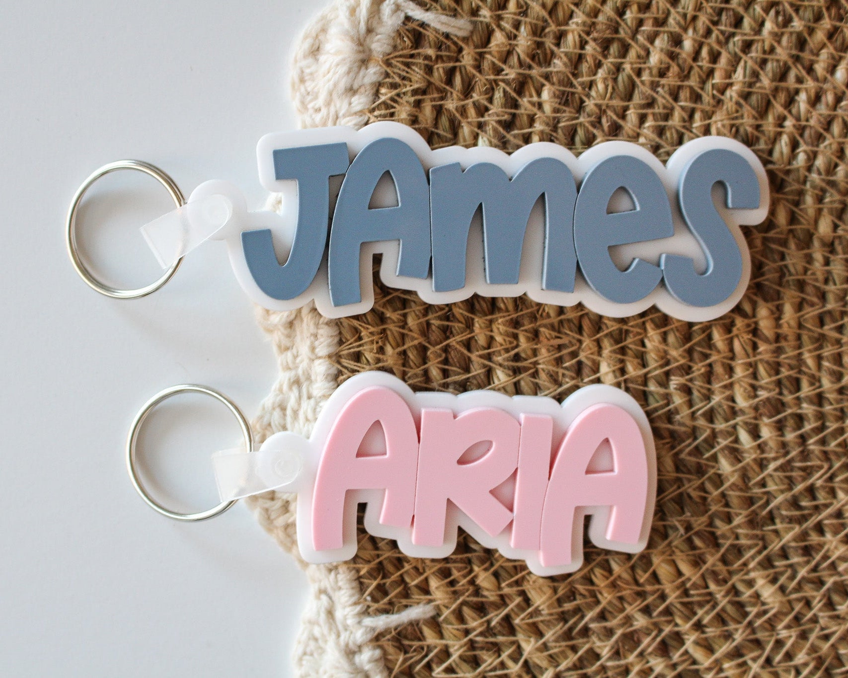 Two personalized layered acrylic name bag tags featuring James in blue and Aria in pink, each with a white backing and silver keyring.