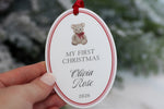Custom baby first Christmas ornament with teddy bear illustration, personalized Olivia Rose name and 2026 date, printed on white oval acrylic with red ribbon.