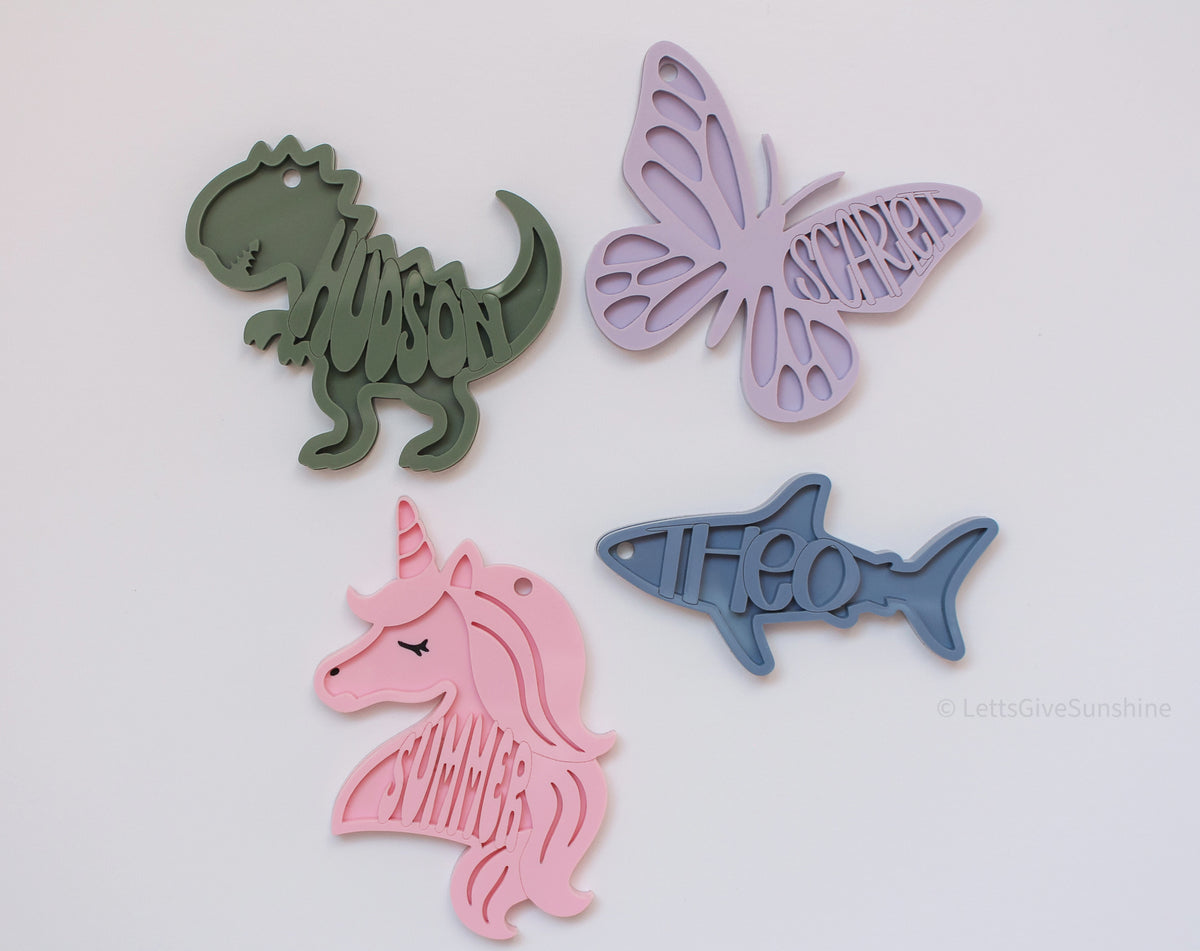 Personalized acrylic bag tags in dinosaur, butterfly, unicorn, and shark designs with the names Tyson, Scarlett, Summer, and Theo.
