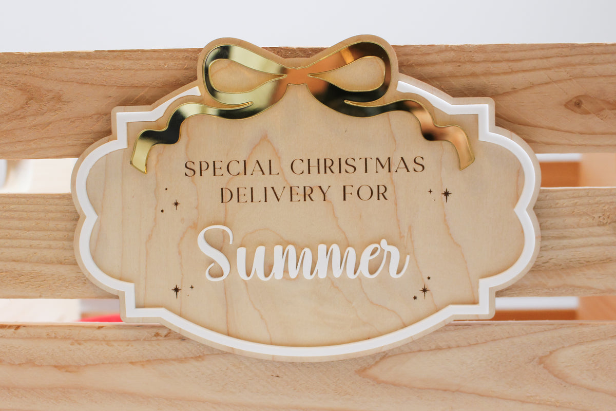 Close-up of a personalized Christmas Eve box sign with natural wood, raised white lettering, a gold mirror acrylic bow and engraved Christmas stars.