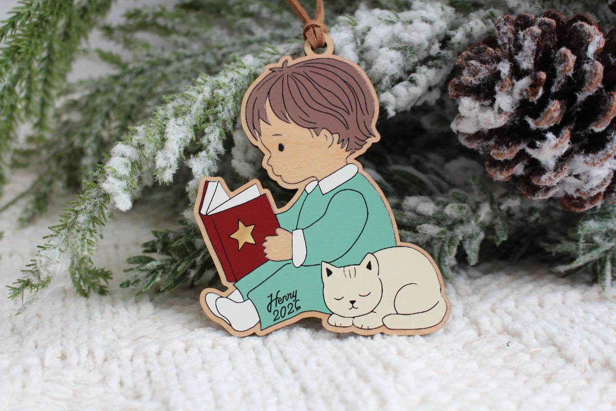 Personalized wooden Christmas ornament featuring a little boy in green pajamas reading a red book beside a sleeping white cat, with the name Henry and year 2026