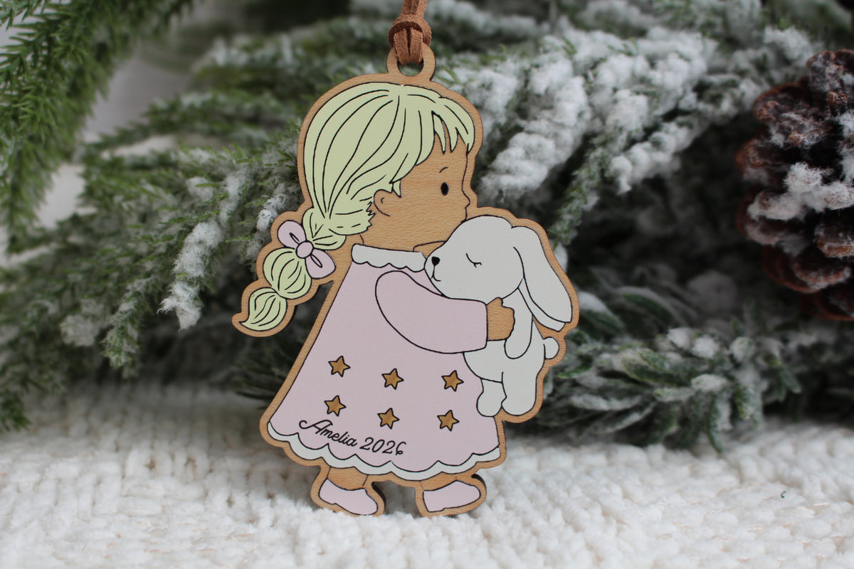 Personalized wooden Christmas ornament featuring a little girl with blonde braids wearing a pink star-patterned nightgown and holding a white bunny, with the name Amelia and year 2026.