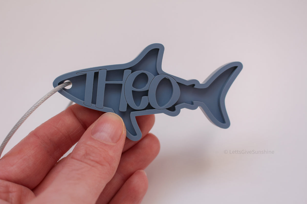 Personalized blue shark-shaped bag tag with the name Theo and a silver cable loop.