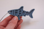 Personalized blue shark-shaped bag tag with the name Theo and a silver cable loop.
