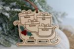Personalized family Christmas ornament 2026 featuring a wooden Santa sleigh filled with engraved name presents, layered holly berries, and a brown hanging ribbon.