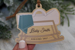 Custom pregnancy announcement Christmas ornament featuring a wooden rocking baby cradle with a blanket, personalized last name, and Arriving year, perfect for a holiday pregnancy reveal.