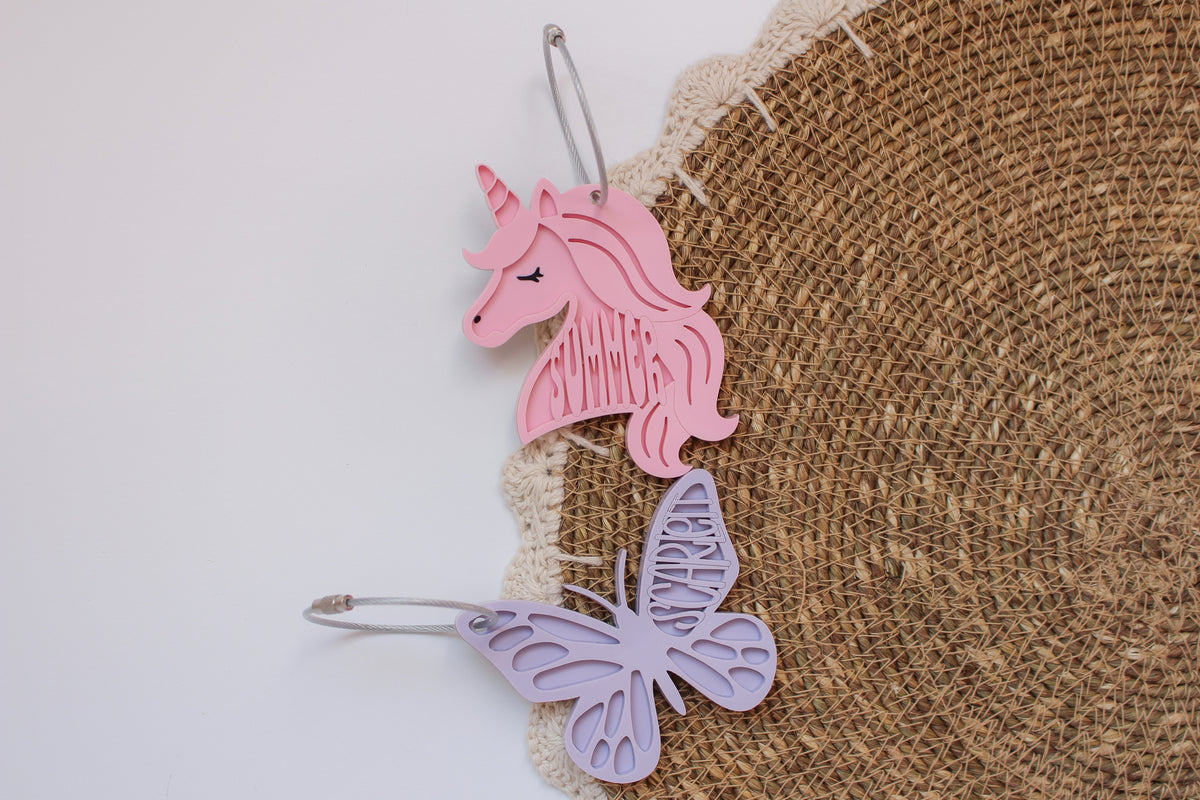 Personalized pink unicorn and lavender butterfly acrylic bag tags with the names Summer and Scarlett, attached to a woven backpack.