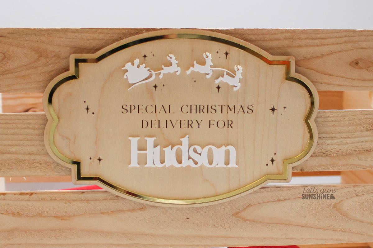 Personalized wooden Christmas Eve box plaque displayed on a wooden crate, featuring the name Hudson in white acrylic, a gold mirror border, Santa's sleigh and reindeer, and engraved stars.