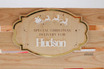 Personalized wooden Christmas Eve box plaque displayed on a wooden crate, featuring the name Hudson in white acrylic, a gold mirror border, Santa's sleigh and reindeer, and engraved stars.