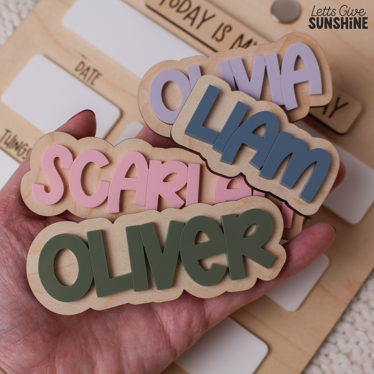 Close-up of interchangeable personalized wooden name pieces with raised acrylic lettering in pink, blue, purple and green, designed for a reusable first day of school dry erase milestone board.