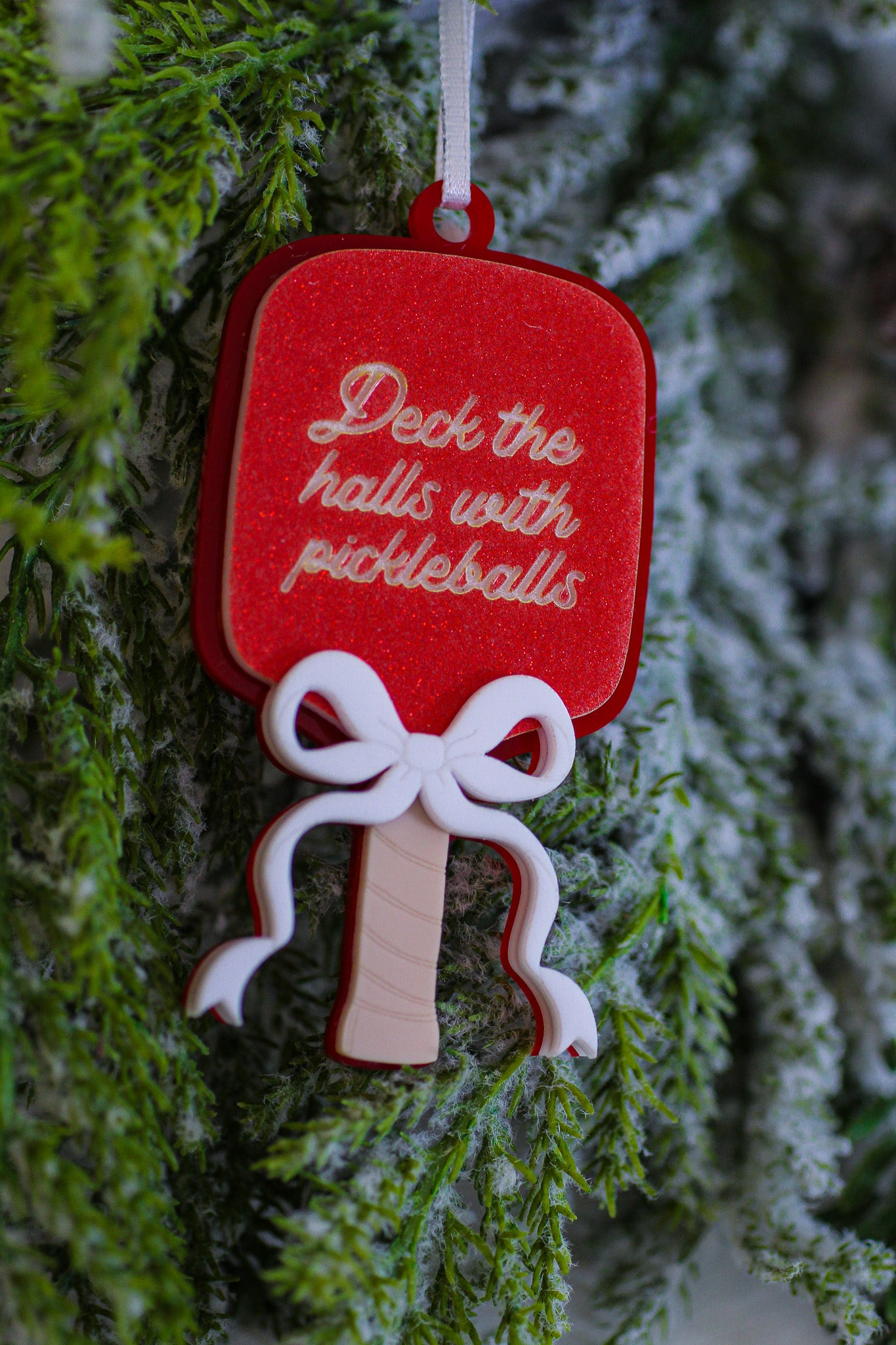 Red glitter pickleball paddle Christmas ornament with a layered white bow and the words Deck the halls with pickleballs.