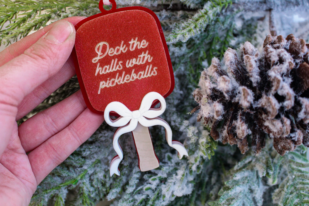 Red glitter pickleball paddle Christmas ornament with a layered white bow and the words Deck the halls with pickleballs.