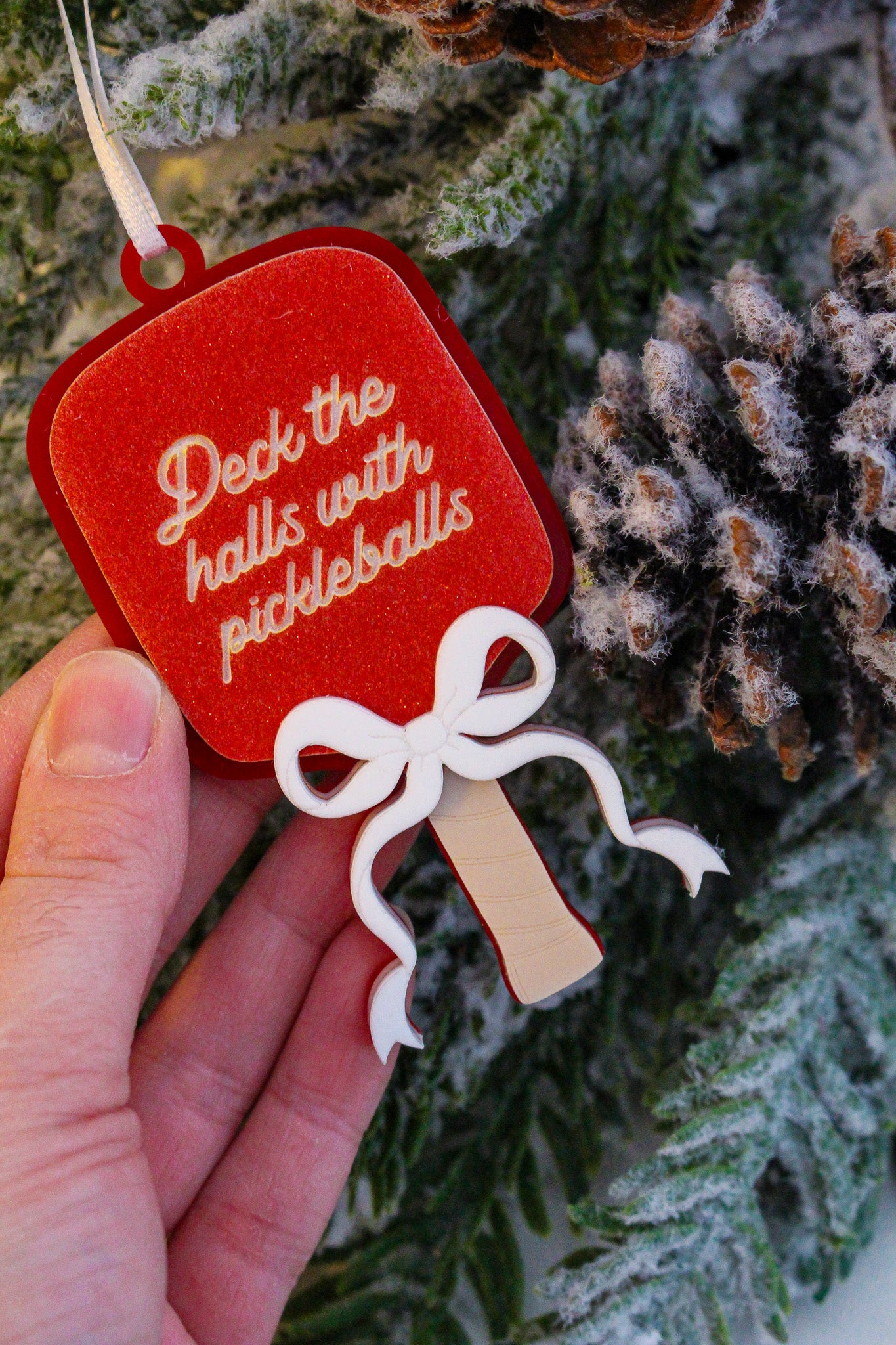 Red glitter pickleball paddle Christmas ornament with a layered white bow and the words Deck the halls with pickleballs.