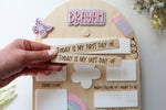 Hand holding interchangeable wooden first day and last day signs in front of a personalized rainbow school board with the name Brayah