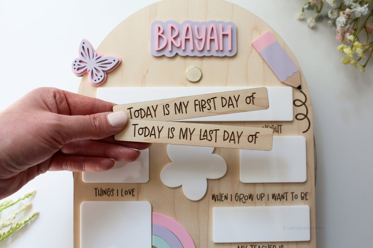 Hand holding interchangeable wooden first day and last day signs in front of a personalized rainbow school board with the name Brayah