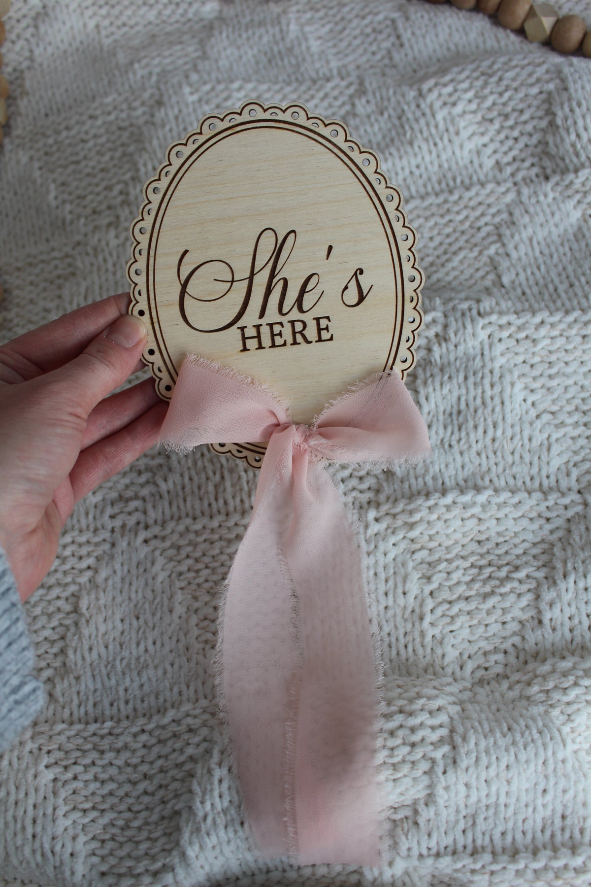 Wooden She’s Here baby announcement sign with pink chiffon bow held over a cozy knit blanket, styled with wooden bead decor.