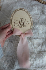 Wooden She’s Here baby announcement sign with pink chiffon bow held over a cozy knit blanket, styled with wooden bead decor.