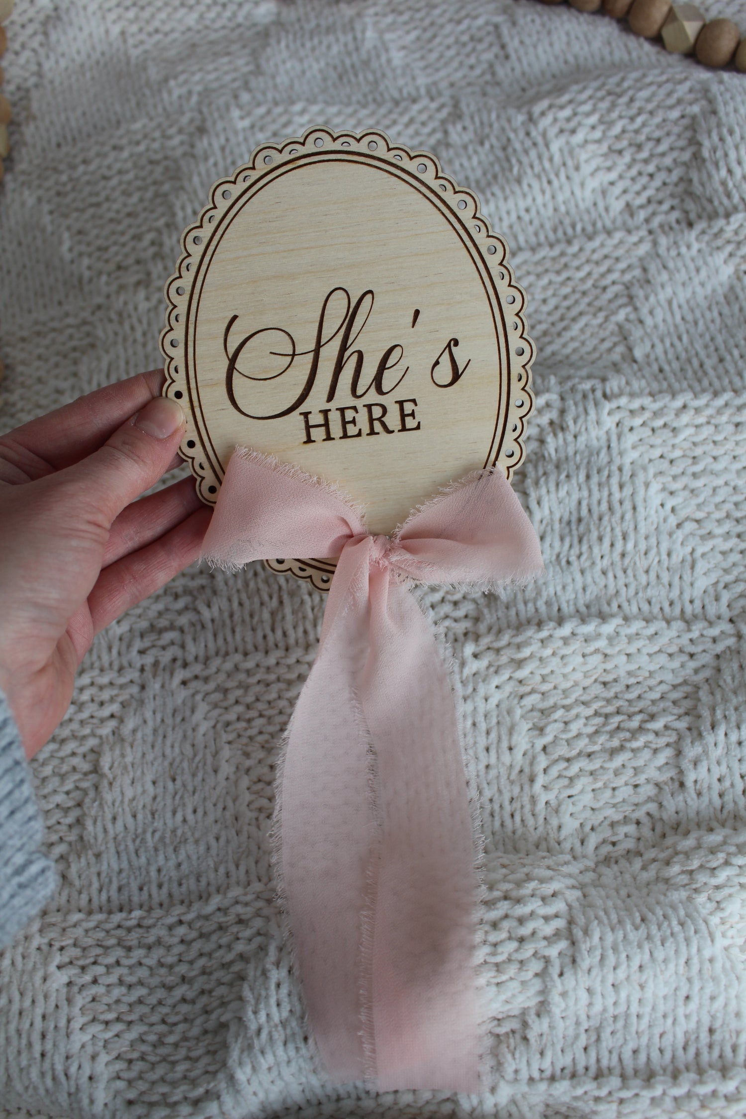 Wooden She’s Here baby announcement sign with pink chiffon bow held over a cozy knit blanket, styled with wooden bead decor.