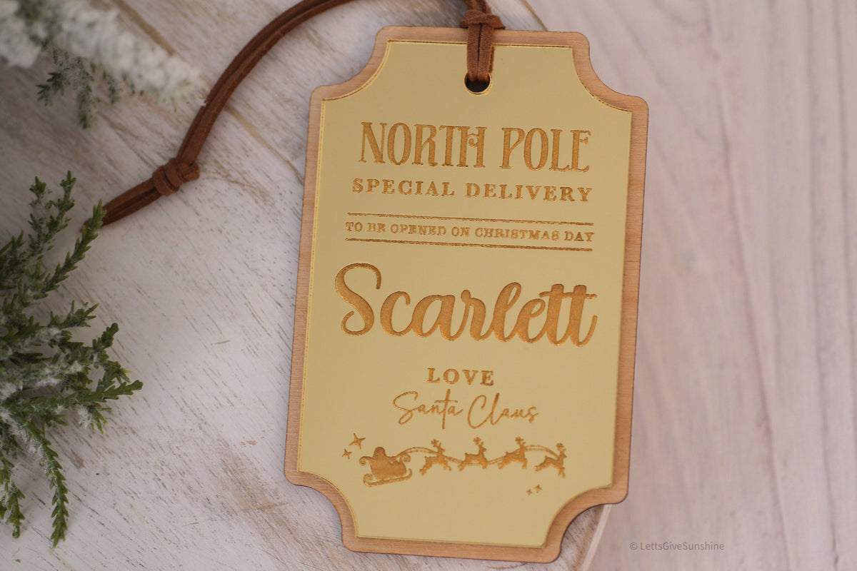 Personalized North Pole Special Delivery Christmas gift tag with Scarlett's name, gold mirrored acrylic, Santa's sleigh and reindeer, and brown hanging cord.