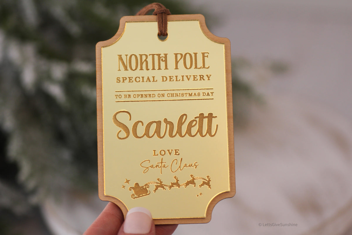 Personalized gold North Pole Christmas gift label with Scarlett's name, special delivery wording, Santa's sleigh illustration and brown faux suede cord.