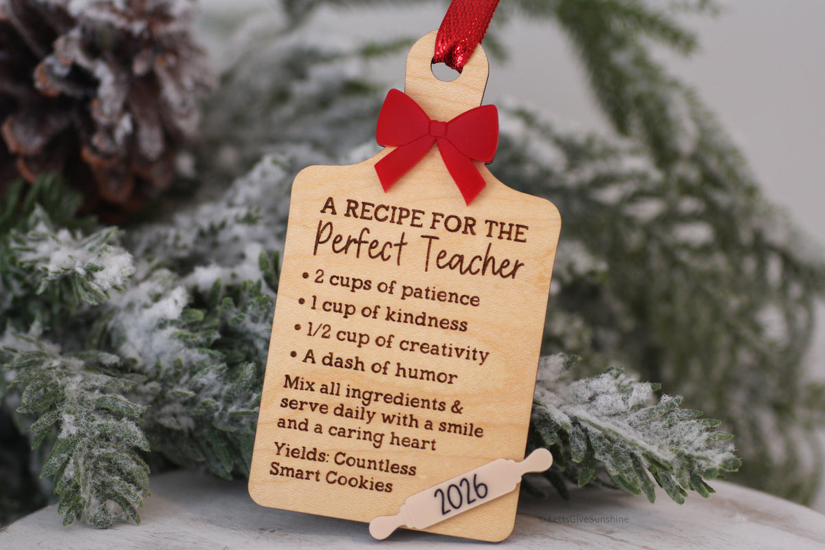 Wooden teacher Christmas ornament shaped like a cutting board with Recipe for the Perfect Teacher engraving, red bow, and 2026 rolling pin detail.