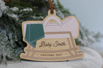 Personalized pregnancy announcement Christmas ornament featuring a wooden rocking baby cradle with a green blanket, custom Baby Smith name, and Arriving 2027, perfect for a holiday pregnancy reveal.