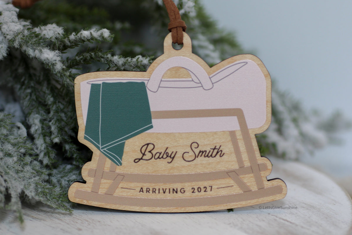 Personalized pregnancy announcement Christmas ornament featuring a wooden rocking baby cradle with a green blanket, custom Baby Smith name, and Arriving 2027, perfect for a holiday pregnancy reveal.